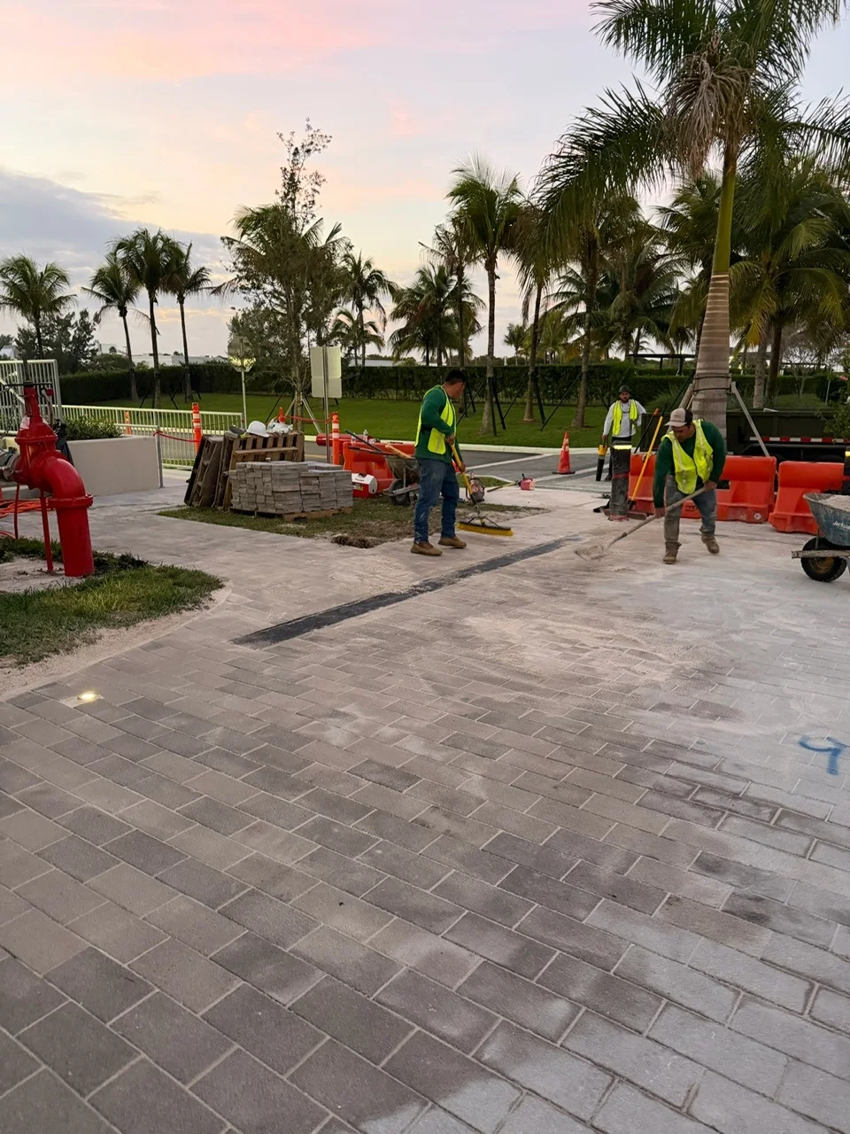 Crew sweeping joint sand across new plaza pavers at sunset beside a row of palms, Miami, FL
