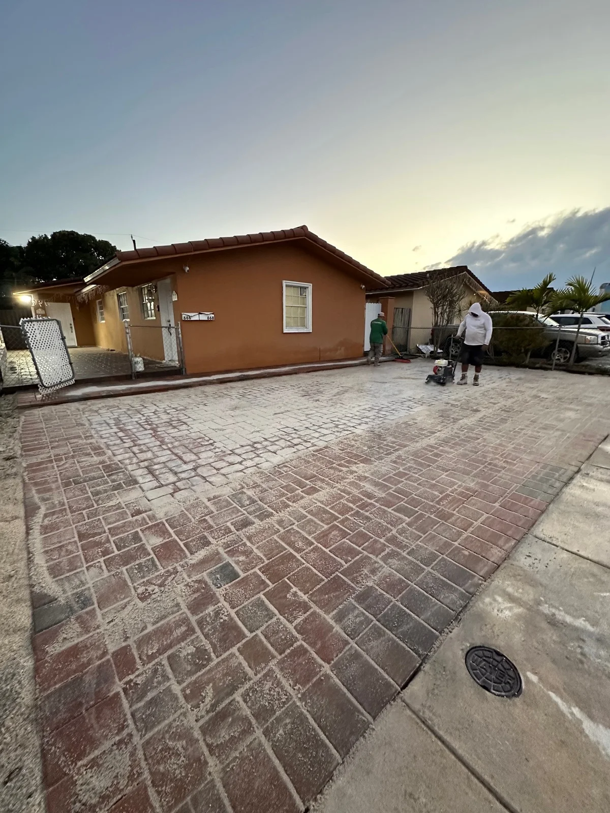 Red-blend paver driveway being compacted at dusk in front of an orange house, Miami, FL