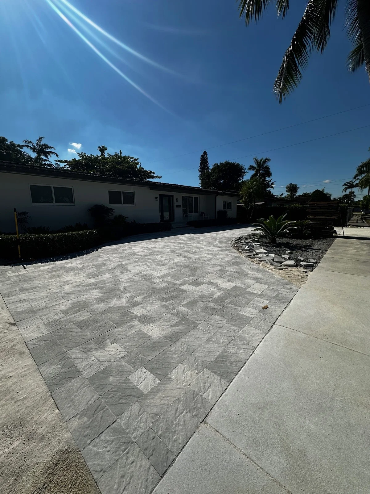 Grey paver driveway and river-rock bed in front of a white house in bright sun, Miami, FL